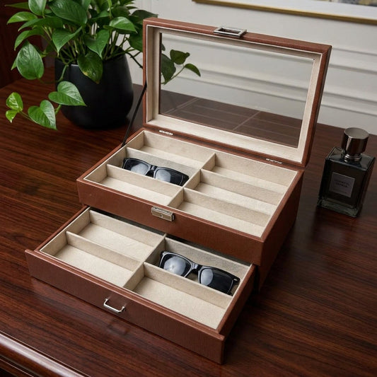 Brown eyeglass case with sunglasses on a wooden surface, next to a plant and perfume bottle.