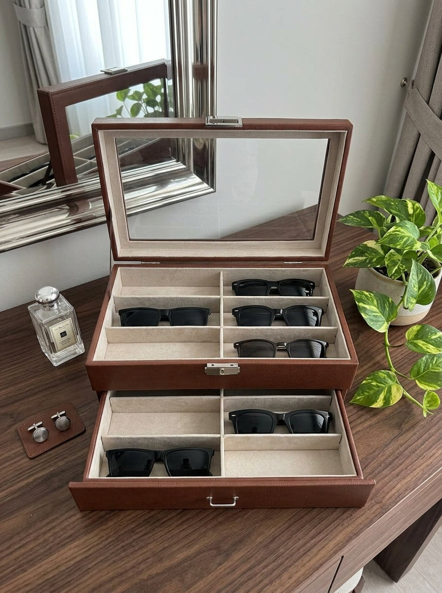 Brown 12-slot double-layer PU leather sunglasses organizer box with clear glass lid open, showing 12 pairs of sunglasses and eyeglasses on a wooden dresser
