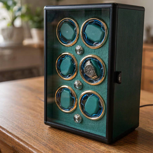 A 6-watch automatic winder box with a dark green textured exterior, clear glass front, and gold-rimmed teal watch slots, sitting on a wooden table.